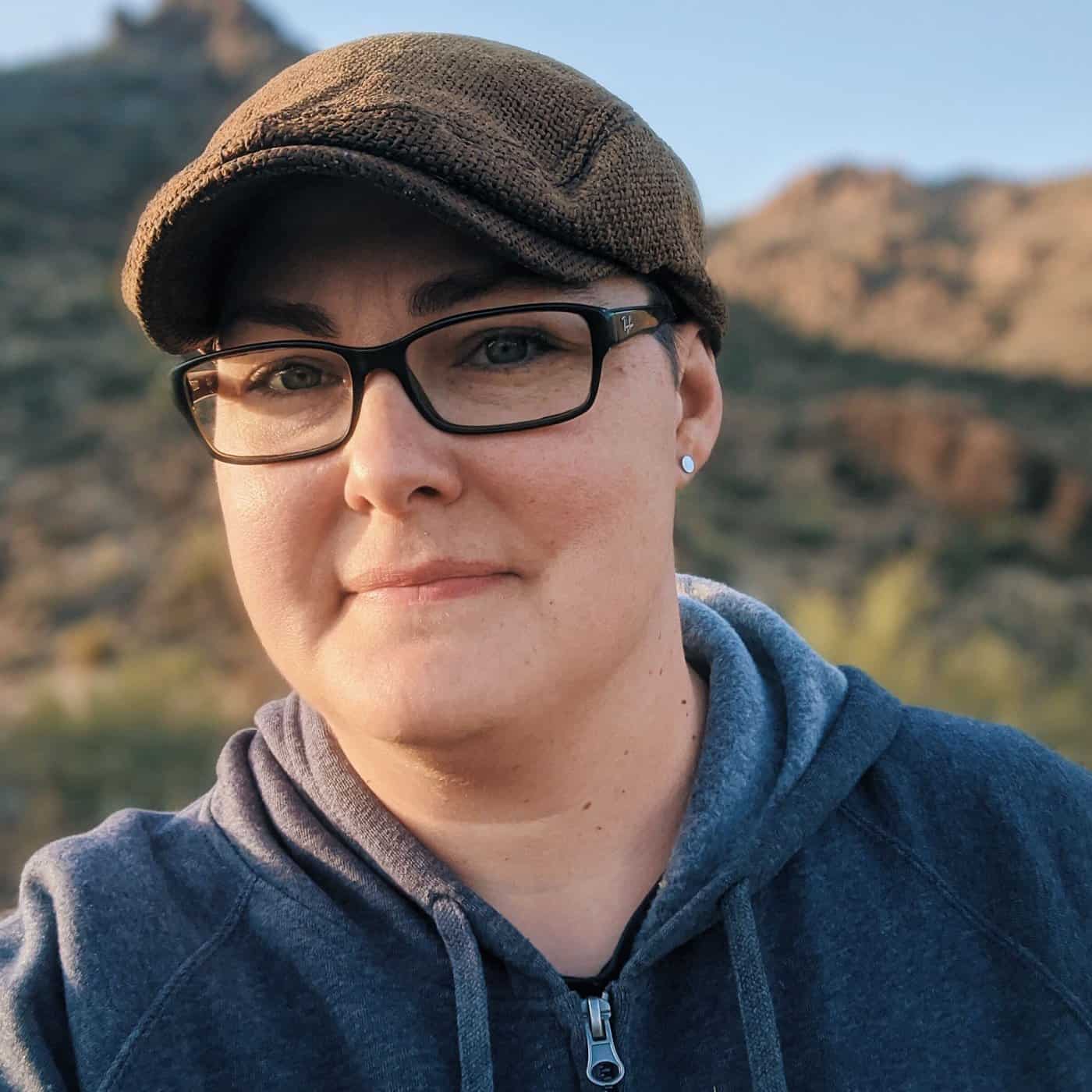 A person wearing a cap and glasses, slightly smiling, with a desert landscape behind.